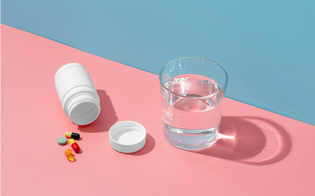 Белая баночка для таблеток, разноцветные капсулы и стакан воды на розовом и голубом фоне